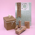 Un coffret cadeau comprenant le Stempel Herz d'Artoz Papier AG, des tampons floraux en bois, un sac Happy Birthday vert menthe, des nœuds en ficelle blancs et une boîte marron, le tout disposé avec art sur un fond rose.
