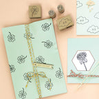 Cadeau emballé dans du papier bleu clair avec des motifs Artoz Papier AG Stempel Kleeblatt, une ficelle jaune et blanche, une étiquette "joyeux anniversaire". À proximité : cartes sur le thème des nuages, Holzstempel Bastelzubehör, timbres et dessin d'hortensia sur papier bleu.