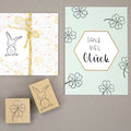 Une carte portant le texte allemand "Ganz viel Glück" et des trèfles dessinés à la main, ainsi que le Artoz Papier AG Stempel Kleeblatt, se trouve à côté d'un cadeau emballé dans du papier blanc et doré avec une ficelle jaune - parfait pour le Bastelzubehör.