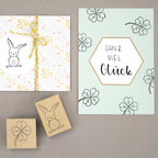 Une carte portant le texte allemand "Ganz viel Glück" et des trèfles dessinés à la main, ainsi que le Artoz Papier AG Stempel Kleeblatt, se trouve à côté d'un cadeau emballé dans du papier blanc et doré avec une ficelle jaune - parfait pour le Bastelzubehör.