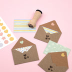 Drei braune Umschläge mit Artoz Papier AG Stempel "kleine Herzen" und gemusterten Karten befinden sich auf einer rosafarbenen Fläche, neben einem Holzstempel, Stern- und Kreisaufklebern und einer gestreiften Notizkarte - ideal zum Basteln.