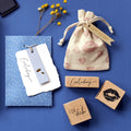 Flat lay sur fond bleu présentant un sac cadeau en tissu, Artoz Papier AG Stempel Kussmund, une enveloppe bleue, une carte blanche, des fleurs jaunes séchées et une étiquette Happy Birthday - des accents idéaux pour les travaux créatifs de Bastelarbeiten.