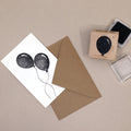 Une enveloppe brune avec une carte présentant deux motifs de ballons noirs, à côté de l'Artoz Papier AG Stempel Luftballon et de deux tampons encreurs sur une surface beige - idéal pour le bricolage créatif.