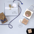 Une présentation à plat de cartes d'anniversaire Artoz Papier AG, d'étiquettes cadeaux Bastelarbeiten, d'un cadeau avec un ruban tressé bleu et jaune, Stempel "mit Liebe selbstgemacht", Holzstempel avec texte allemand, et d'un sac cadeau blanc festonné sur gris clair.