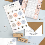 Une mise en page plate de papeterie pour Bastelarbeiten, avec des autocollants de fleurs et d'abeilles, des papiers à motifs, Artoz Papier AG Stempel Pflanze, et des enveloppes avec des noms écrits à la main et des motifs d'abeilles sur fond beige - idéal pour des Einladungskarten créatives.