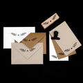 Un ensemble d'Einladungskarten et d'enveloppes en blanc, marron et beige, chacune représentant une simple branche feuillue de l'Artoz Papier AG Stempel Pflanze. Une pince en métal et un fond noir complètent cette élégante scène de Bastelarbeiten.
