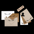 Un ensemble d'Einladungskarten et d'enveloppes en blanc, marron et beige, chacune représentant une simple branche feuillue de l'Artoz Papier AG Stempel Pflanze. Une pince en métal et un fond noir complètent cette élégante scène de Bastelarbeiten.