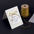Une carte faite à la main avec des feuilles d'or, un nœud doré et la mention "schön dass du da bist" d'Artoz Papier AG. À côté, une bobine de ficelle dorée et du papier artisanal décoré sur une surface noire.