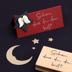 Une carte estampillée du stempel "schön dass du da bist" d'Artoz Papier AG repose sur une surface sombre, entourée de découpes Bastelarbeiten en forme de lune et d'étoile, d'un Holzstempel et d'une étoile noire décorée d'un ange.