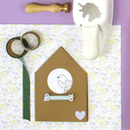 Eine flache Anordnung von Bastelmaterial auf Lila, mit Blumenpapier, Washi-Tape, grünem Band, einer Einhorn-Stanze, einer handgemachten Geburtstagskarte in Form eines Hauses mit dem Stempel Vogel von Artoz Papier AG und Blumendekor für Bastelarbeiten.