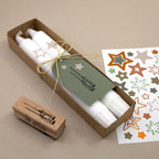 Un lot de deux bougies blanches « Stempel Kerzen » de la marque Artoz Papier AG, présentées avec une étiquette verte nouée par un cordon doré, comprend un tampon en forme d'étoile, idéal pour les loisirs créatifs, ainsi qu'une feuille d'autocollants colorés en forme d'étoile, le tout disposé sur un support beige.