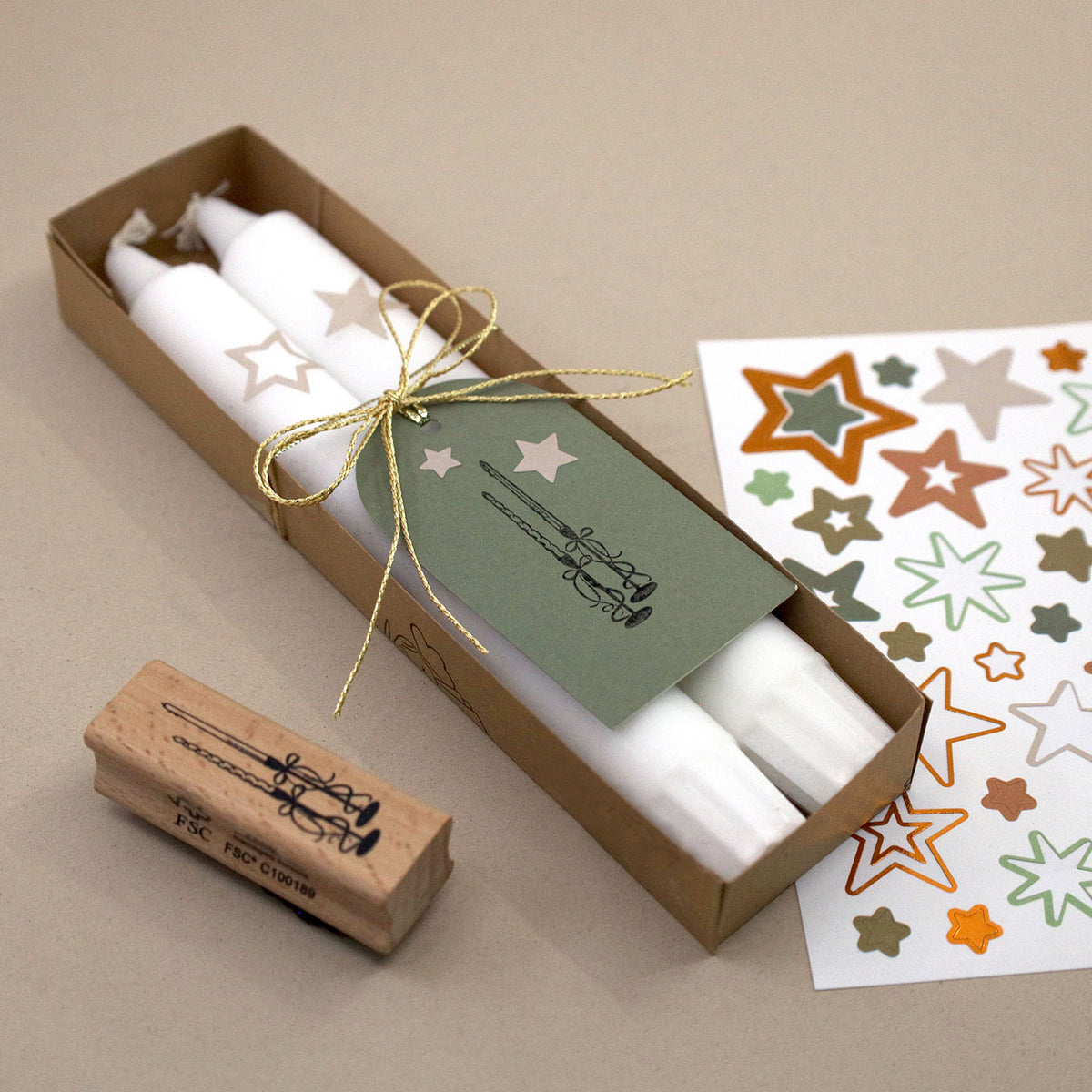Un lot de deux bougies blanches « Stempel Kerzen » de la marque Artoz Papier AG, présentées avec une étiquette verte nouée par un cordon doré, comprend un tampon en forme d'étoile, idéal pour les loisirs créatifs, ainsi qu'une feuille d'autocollants colorés en forme d'étoile, le tout disposé sur un support beige.
