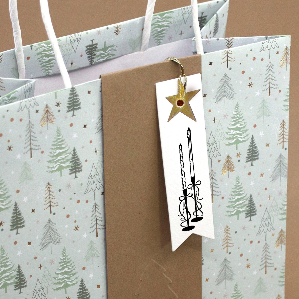Un sac cadeau orné de motifs de sapins, décoré à l'aide du tampon « Sapin » d'Artoz Papier AG, ainsi qu'une étiquette cadeau blanche, ornée d'un motif de bougie réalisé à l'aide du tampon « Bougies » d'Artoz Papier AG – idéal pour vos projets de bricolage ou pour personnaliser vos cadeaux avec votre tampon en bois préféré « Hêtre ».