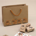 Un sac cadeau marron aux anses orange, orné de motifs de citrouilles estampés à l'aide du tampon « Stempel Kürbis » d'Artoz Papier AG, est idéal pour les projets de bricolage. À proximité, on trouve des tampons en forme de citrouille, des autocollants en forme d'étoile, des tampons en bois de hêtre et des tampons encreurs, le tout posé sur un support beige.