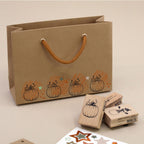 Un sac cadeau marron aux anses orange, orné de motifs de citrouilles estampés à l'aide du tampon « Stempel Kürbis » d'Artoz Papier AG, est idéal pour les projets de bricolage. À proximité, on trouve des tampons en forme de citrouille, des autocollants en forme d'étoile, des tampons en bois de hêtre et des tampons encreurs, le tout posé sur un support beige.