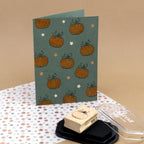Une carte de vœux verte, de format vertical, est ornée de motifs de citrouilles orange et de petites étoiles. Devant elle se trouvent le tampon « Stempel Kürbis » d’Artoz Papier AG, un tampon encreur noir et du papier à motifs floraux : une source d’inspiration idéale pour votre prochain projet de bricolage. Le fond beige clair met en valeur la polyvalence créative des tampons en bois, ce qui permet de réaliser facilement des motifs festifs tels que le tampon « Stempel Tannenbaum ».