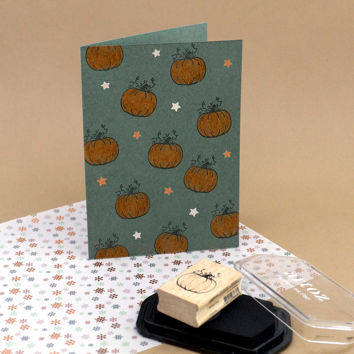 Une carte de vœux verte, de format vertical, est ornée de motifs de citrouilles orange et de petites étoiles. Devant elle se trouvent le tampon « Stempel Kürbis » d’Artoz Papier AG, un tampon encreur noir et du papier à motifs floraux : une source d’inspiration idéale pour votre prochain projet de bricolage. Le fond beige clair met en valeur la polyvalence créative des tampons en bois, ce qui permet de réaliser facilement des motifs festifs tels que le tampon « Stempel Tannenbaum ».