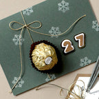 Eine grüne Karte mit einem Schneeflockenmuster, das mit dem Stempel „Schneeflocke klein“ von Artoz Papier AG gestaltet wurde, ist mit einer goldenen Schnur zusammengebunden und steht auf einem Tisch neben einer in Goldpapier verpackten Praline und nummerförmigen Keksen, die zusammen die Zahl 27 bilden – alles wunderschön arrangiert.