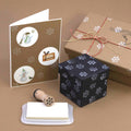 Auf einer braunen Unterlage sind eine Grußkarte, zwei Geschenkboxen und ein Stempel mit Stempelkissen angeordnet; alle Artikel sind mit weißen Schneeflockenmotiven verziert. Diese bezaubernden Stücke eignen sich perfekt für DIY-Bastelprojekte und lassen sich mit Ihrem Lieblings-Holzstempel oder dem Stempel „Schneeflocke klein“ von Artoz Papier AG individuell gestalten, um ihnen eine festliche Note zu verleihen.