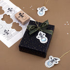 Eine schwarze Geschenkbox mit goldenen Punkten, die mit einem grünen Band verziert ist, steht auf einer braunen Unterlage. Daneben liegen Geschenkanhänger in Form von Schneemännern aus bedrucktem Papier sowie der „Stempel Schneemann“ von Artoz Papier AG – ideal, um Ihren Scrapbook-Projekten oder Weihnachtsgeschenken eine kreative Note zu verleihen.