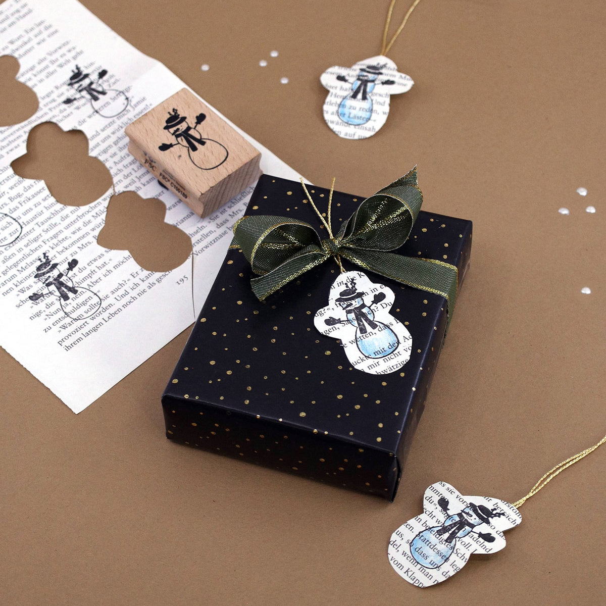 Eine schwarze Geschenkbox mit goldenen Punkten, die mit einem grünen Band verziert ist, steht auf einer braunen Unterlage. Daneben liegen Geschenkanhänger in Form von Schneemännern aus bedrucktem Papier sowie der „Stempel Schneemann“ von Artoz Papier AG – ideal, um Ihren Scrapbook-Projekten oder Weihnachtsgeschenken eine kreative Note zu verleihen.