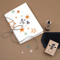 Eine handgefertigte Weihnachtskarte mit Sternstempeln und dem Stempel „Schneemann“ von Artoz Papier AG, die mit einer Schnur zusammengebunden ist, liegt auf einem braunen Untergrund. Daneben liegen ein Stempelkissen, ein Stempel „Tannenbaum“ und einige sternförmige Ausschnitte – idealer Bastelbedarf für Ihre festlichen Kreationen.
