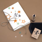 Eine handgefertigte Weihnachtskarte mit Sternstempeln und dem Stempel „Schneemann“ von Artoz Papier AG, die mit einer Schnur zusammengebunden ist, liegt auf einem braunen Untergrund. Daneben liegen ein Stempelkissen, ein Stempel „Tannenbaum“ und einige sternförmige Ausschnitte – idealer Bastelbedarf für Ihre festlichen Kreationen.