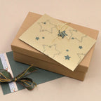 Auf einer beigen Unterlage steht eine braune Geschenkbox mit einer Karte mit goldenem Sternenmuster, verziert mit dem „Stempel Stern“ von Artoz Papier AG und mit einer Schnur zusammengebunden. Unter der Box liegt ein grüner Umschlag, der mit einem Zierband verziert ist – eine ideale Wahl für Bastelarbeiten und kreative Weihnachtsgeschenke.