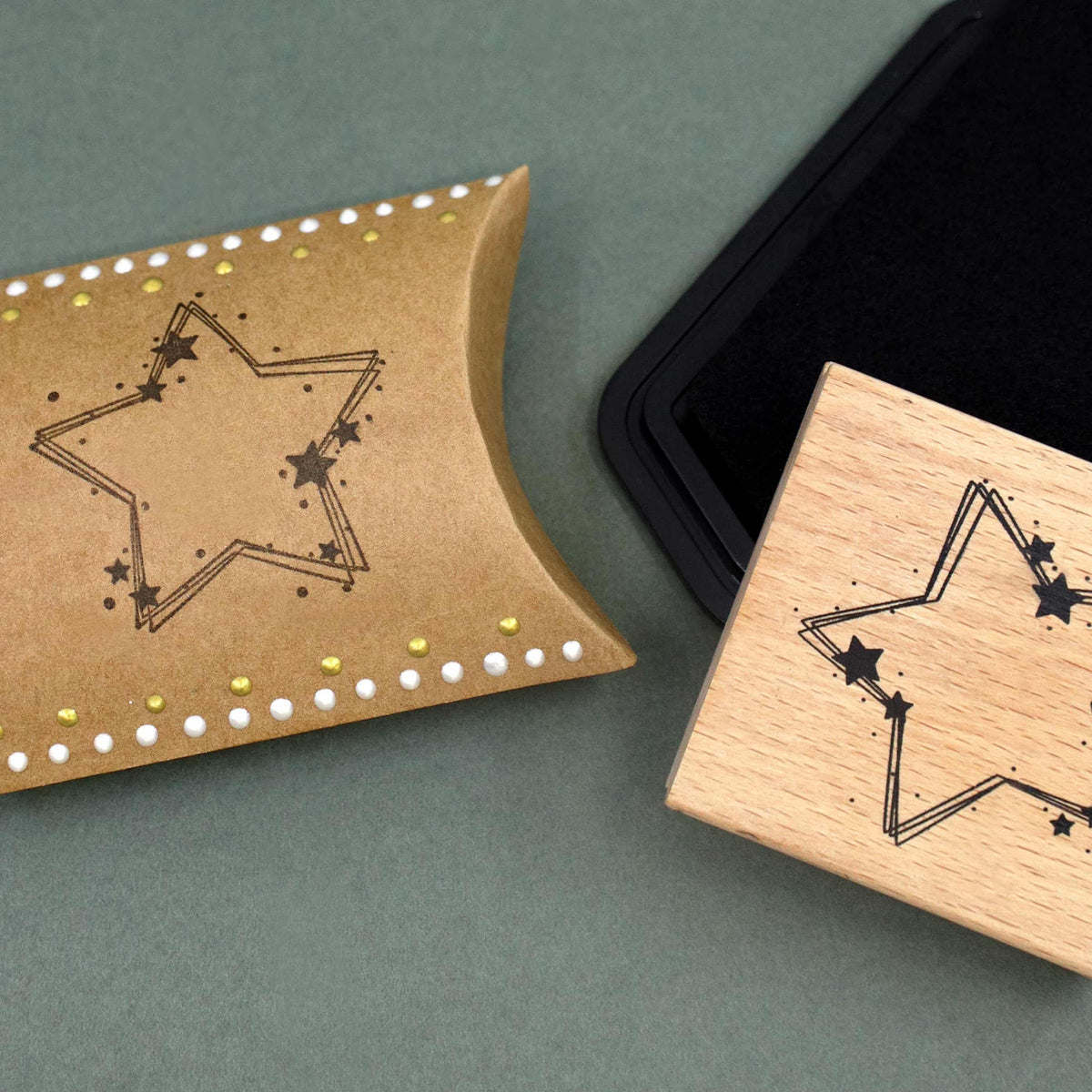 Auf einer grünen Fläche steht eine braune Pappschachtel mit dem Motiv „Stempel Stern dekoriert“ von Artoz Papier AG neben einem Holzstempel mit dem passenden Sternmotiv – ideal für Bastelbedarf. In der Ecke ist teilweise ein schwarzes Stempelkissen zu sehen.
