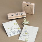Deux cartes décorées portant les inscriptions « Nima » et « Leon », ornées de timbres sur le thème des étoiles et des fêtes, sont exposées à proximité de deux tampons DIY de la marque Artoz Papier AG Holzstempel — dont un « Stempel Weihnachtskugel » représentant le contour d’une boule de Noël — et d’un petit trombone en métal, le tout disposé sur un support marron.