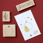 Une carte blanche ornée d'une boule de Noël dorée et d'étoiles est entourée de trois tampons en bois « Bastel Stempel » de la marque Artoz Papier AG, aux motifs festifs, dont le « Stempel Weihnachtskugel », le tout disposé sur un fond rouge vif.