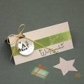 Une carte beige ornée du tampon « Stempel Winterzauber » d'Artoz Papier AG est agrémentée d'un ruban vert, d'un cordon doré et d'une décoration représentant un chalet d'hiver. Parmi les autres éléments décoratifs, on trouve une étoile rose, une petite illustration représentant un cadeau vert, ainsi que de délicats détails de scrapbooking aux motifs festifs de sapins de Noël, qui ajoutent une touche de charme supplémentaire à cette carte de saison.