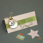 Une carte beige ornée du tampon « Stempel Winterzauber » d'Artoz Papier AG est agrémentée d'un ruban vert, d'un cordon doré et d'une décoration représentant un chalet d'hiver. Parmi les autres éléments décoratifs, on trouve une étoile rose, une petite illustration représentant un cadeau vert, ainsi que de délicats détails de scrapbooking aux motifs festifs de sapins de Noël, qui ajoutent une touche de charme supplémentaire à cette carte de saison.