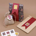 Un sac cadeau à motifs floraux, un sac en toile de jute muni d’une étiquette et une enveloppe marron ornée d’un ruban rouge — tous trois décorés de petits autocollants « Artoz Sticker Advent 1-24 in Briefmarkenform » de la marque Artoz Papier AG — sont disposés sur un fond marron. Une feuille d’autocollants destinée à la décoration des cadeaux se trouve à proximité.