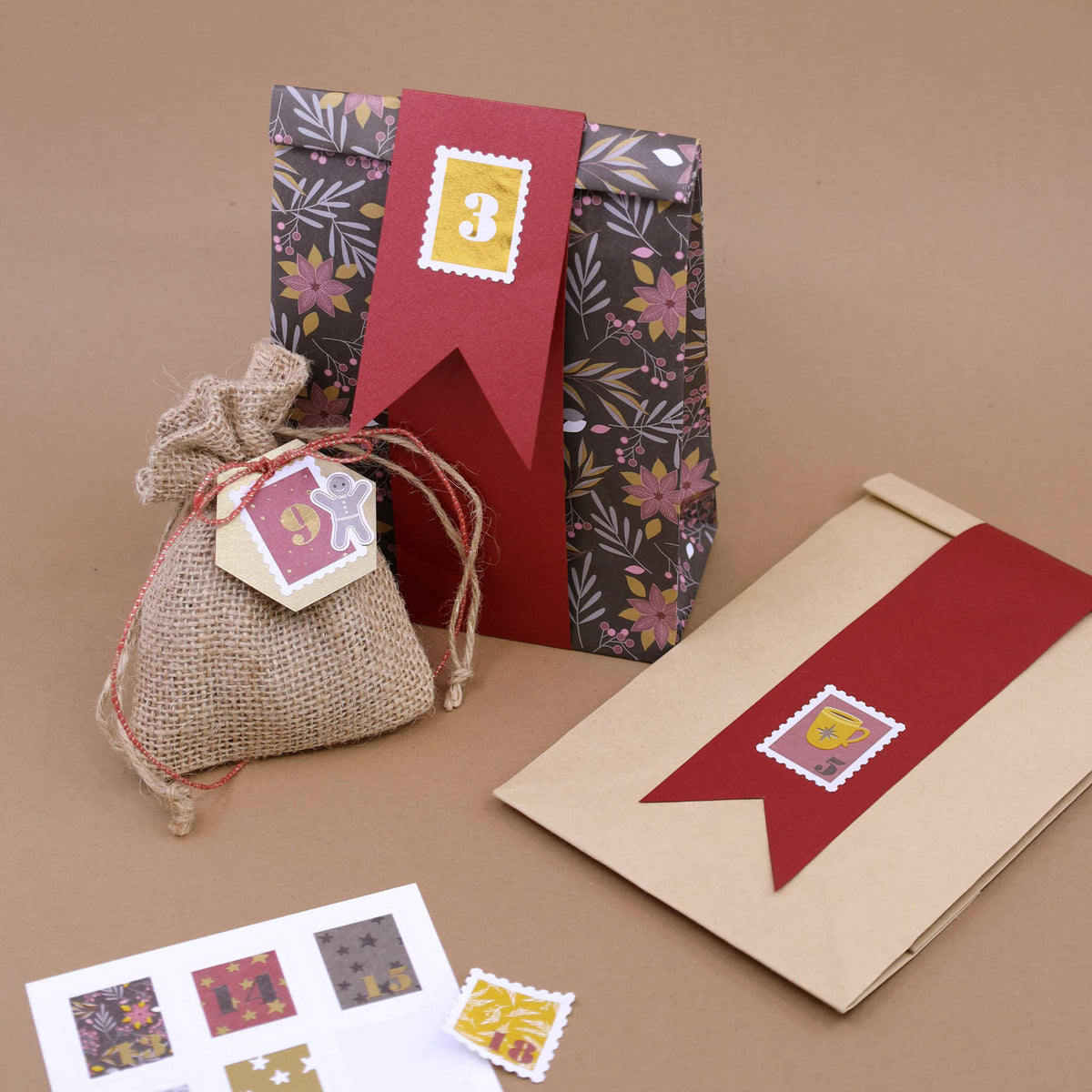 Un sac cadeau à motifs floraux, un sac en toile de jute muni d’une étiquette et une enveloppe marron ornée d’un ruban rouge — tous trois décorés de petits autocollants « Artoz Sticker Advent 1-24 in Briefmarkenform » de la marque Artoz Papier AG — sont disposés sur un fond marron. Une feuille d’autocollants destinée à la décoration des cadeaux se trouve à proximité.