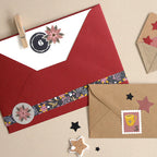 Ein roter Umschlag mit Blumenmuster und ein brauner Umschlag, verziert mit Stern- und Tassen-Aufklebern – darunter die „Artoz Sticker Adventsnummern 1–24 schwarz“ von Artoz Papier AG – sind auf einer beigen Unterlage angeordnet und von schwarzen und goldenen Sternausschnitten umgeben – ideal zum Dekorieren von Geschenken.