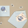 Une enveloppe et une carte bleu clair avec Alex et un cœur sont ornées d'une épingle à fleur bleue. Une autre enveloppe contient une carte fleurie avec un lapin blanc et un autocollant Artoz 3D "Biene mit Blume" d'Artoz Papier AG, ainsi que des autocollants abeilles 3D sur fond beige.