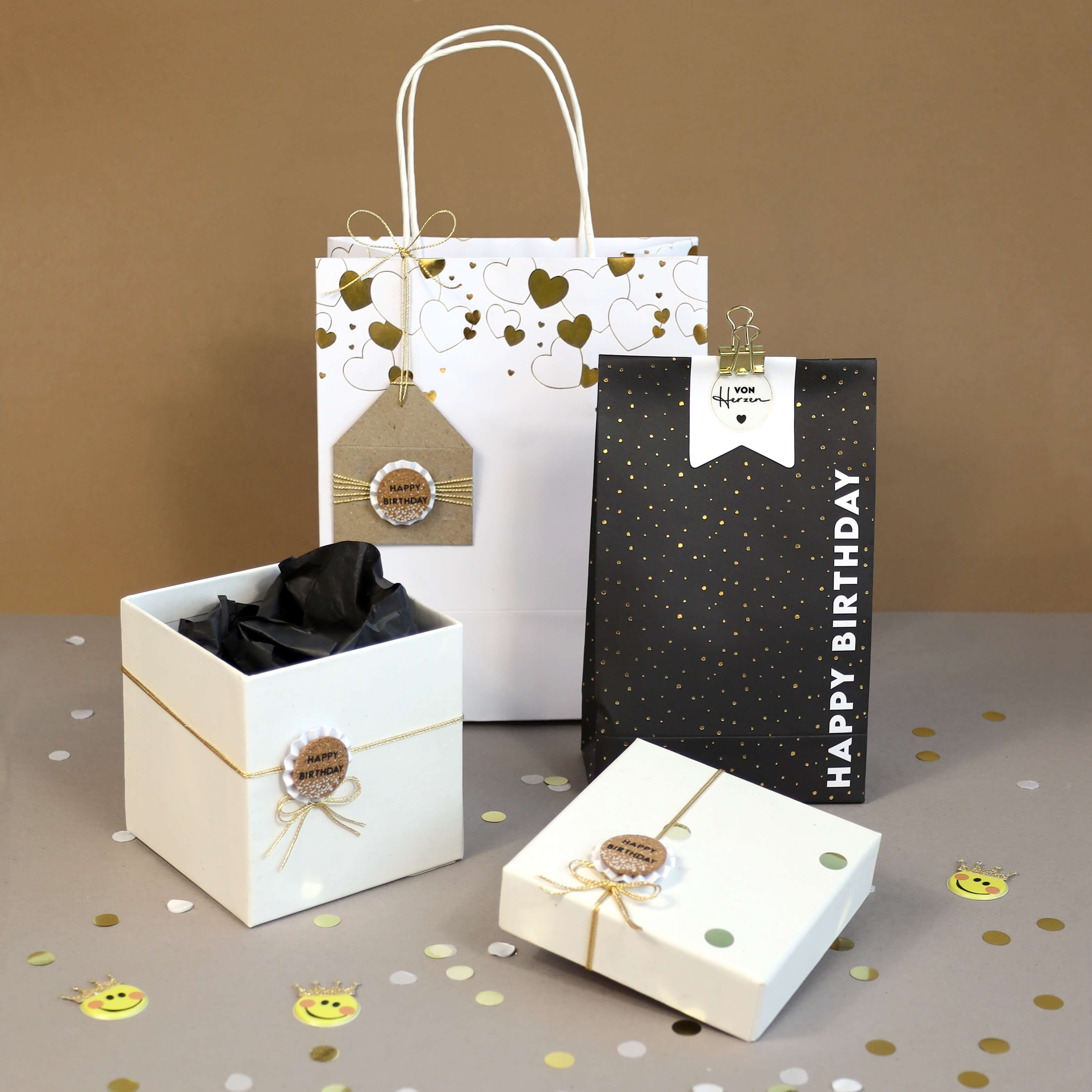 Sacs et boîtes cadeaux avec des accents dorés et noirs, l'autocollant 3D "Happy Birthday" d'Artoz Papier AG, des nœuds en ficelle et des étiquettes sur une surface grise avec des confettis. Un sac porte l'inscription "Happy Birthday" ; les autres ont des étiquettes décoratives. Fond brun neutre.
