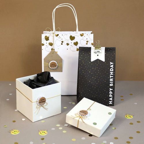 Sacs et boîtes cadeaux avec des accents dorés et noirs, l'autocollant 3D "Happy Birthday" d'Artoz Papier AG, des nœuds en ficelle et des étiquettes sur une surface grise avec des confettis. Un sac porte l'inscription "Happy Birthday" ; les autres ont des étiquettes décoratives. Fond brun neutre.
