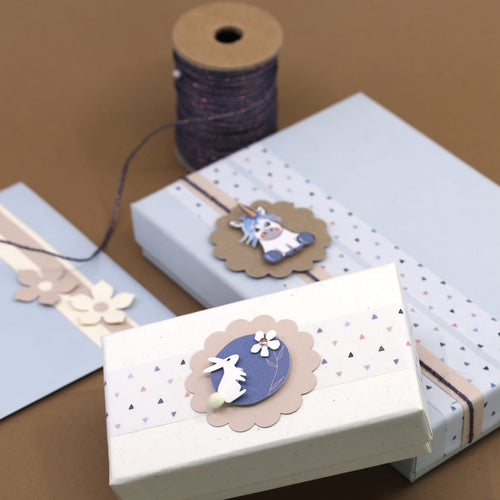 Deux boîtes-cadeaux pastel aux motifs floraux et géométriques sont décorées avec l'autocollant 3D Artoz "Hase mit Blume" d'Artoz Papier AG. Chaque boîte comporte une étiquette en papier festonnée, tandis qu'une bobine de ficelle repose à proximité sur la surface brune.