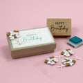 Un coffret cadeau d'anniversaire Artoz Papier AG décoré de l'autocollant 3D Artoz "Herzballone", comprenant une carte "Joyeux anniversaire", des fleurs décoratives en papier, un tampon en bois et un tampon encreur, le tout disposé sur un fond rose.