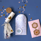 Un layon plat avec des confettis, des Bastelzubehör, un bocal en verre avec couvercle en liège et étiquette Einladung, une enveloppe florale festonnée étiquetée Danke, du papier roulé noué avec Artoz Sticker Herz gold by Artoz Papier AG, et une carte rose avec des autocollants hexagonaux.