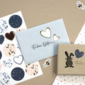 Auf einem blauen Umschlag befinden sich die Artoz Sticker Herzen der Artoz Papier AG mit goldenen Herzen und dem Text "Frohe Ostern", florale 3D-Sticker und ein handgefertigtes Schweizer Geschenk aus braunem Papier mit einem schwarzen Hasen, alles auf einer beigen Oberfläche.