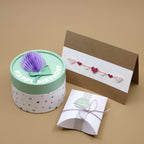 Eine runde Geschenkschachtel mit "Happy Birthday"-Text, lila Dekor, eine braune Karte mit Herzmotiven mit Artoz 3D Sticker "Herzgirlande" von Artoz Papier AG, und eine kleine weiße handgefertigte Schachtel mit Schnur auf beigem Hintergrund.