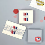 Une mise à plat de trois cadeaux d'anniversaire emballés, deux avec des étiquettes Happy Birthday et de petites bougies, décorés avec Bastelzubehör et Artoz 3D Sticker "Kerzen Happy Birthday" par Artoz Papier AG, entourés de cœurs et de cercles en papier sur fond gris.
