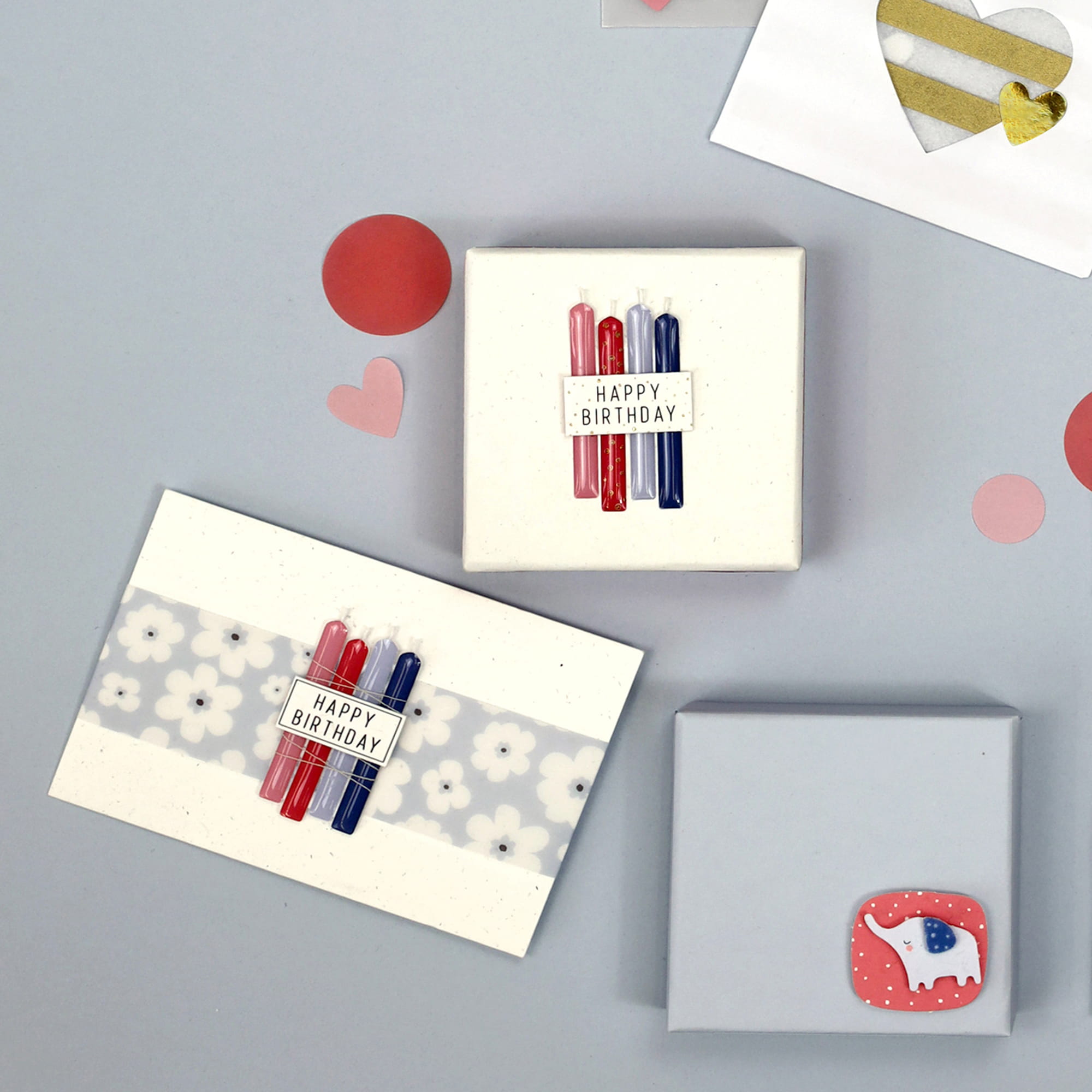 Une mise à plat de trois cadeaux d'anniversaire emballés, deux avec des étiquettes Happy Birthday et de petites bougies, décorés avec Bastelzubehör et Artoz 3D Sticker "Kerzen Happy Birthday" par Artoz Papier AG, entourés de cœurs et de cercles en papier sur fond gris.