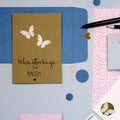 Une carte brune faite à la main avec l'autocollant 3D "Sommervogel" d'Artoz Papier AG et la phrase "Schmetterlinge im Bauch" repose sur une enveloppe bleue, entourée de papier rose à motifs, de crayons, d'un stylo et de papiers découpés.