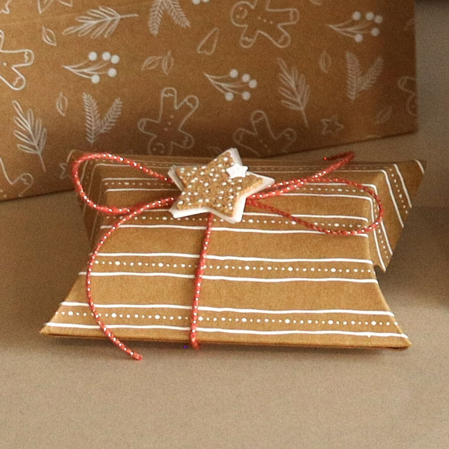 Un petit cadeau en forme d'oreiller, emballé dans du papier kraft à rayures blanches, noué avec une ficelle rouge et agrémenté d'un autocollant 3D « Stern Kork » de la marque Artoz Papier AG, pour une touche de charme typique du design suisse.