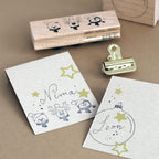 Deux cartes portant les noms « Nima » et « Leon », ornées d'adorables animaux tamponnés coiffés de chapeaux de fête et de bois, d'autocollants scintillants « Artoz Sticker Sternen Gold » de la société Artoz Papier AG, d'une pince dorée, des touches d'autocollants et un tampon en bois sur fond marron – idéales pour vos projets de décoration créatifs à réaliser vous-même pendant la période de Noël.