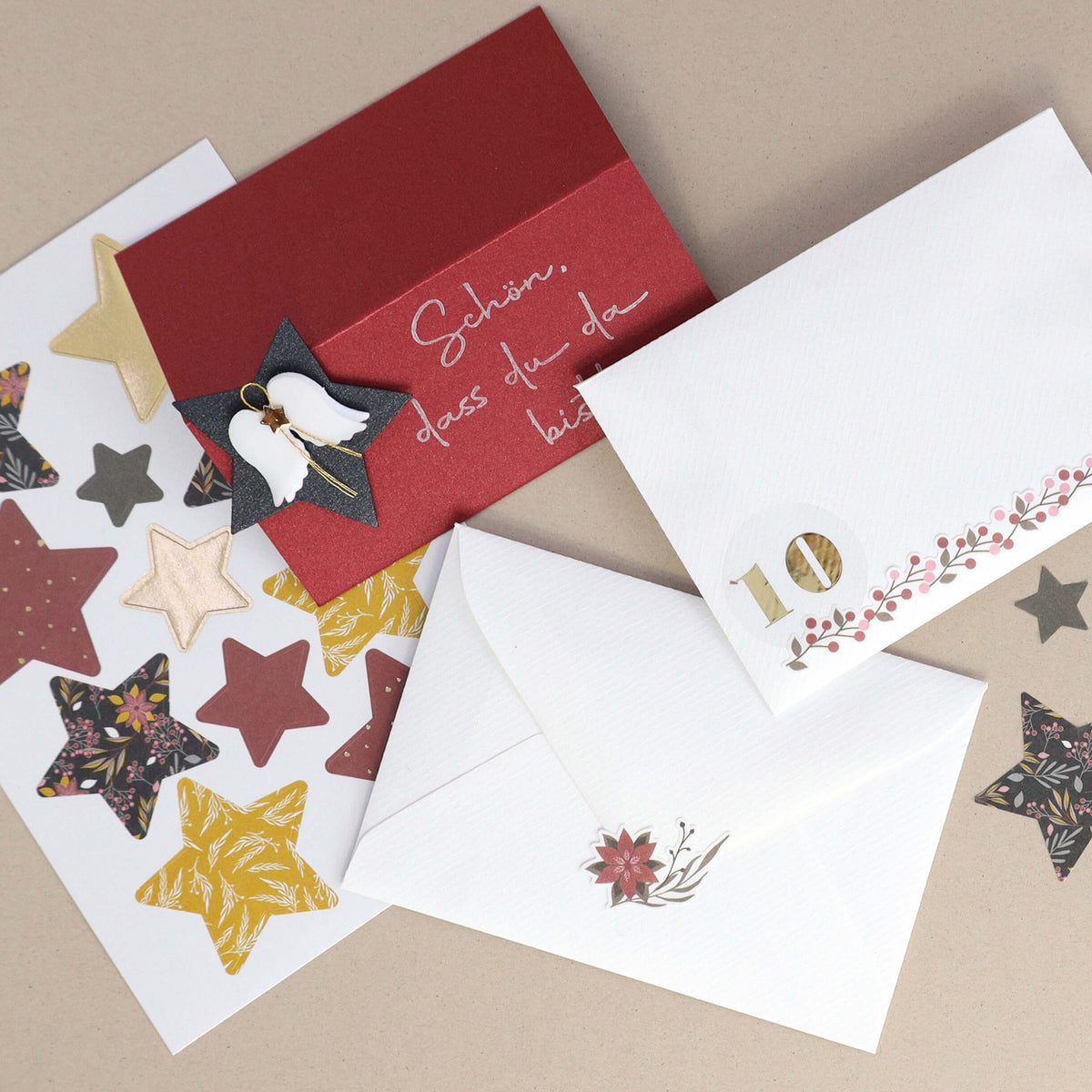 Trois enveloppes — une rouge et deux blanches — sont ornées d'étoiles, d'un ange en papier et de motifs floraux. Les autocollants « Artoz Sticker Sternen Diverse Farben » de la société Artoz Papier AG ainsi qu'une petite décoration en forme de « 10 » viennent rehausser cette composition décorative à réaliser soi-même sur un fond beige.